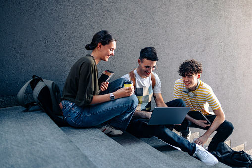 Close up shot of a group of young happy students sitting on the stairs, smiling while chatting to each other. The man in the middle is using the laptop computer while the other man and a woman are looking at what he is showing them