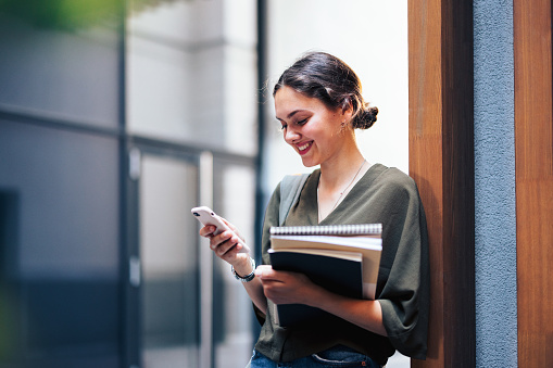 Portrait of a beautiful happy  young female student leaning against a wall with wooden strip cladding. She is holding her notebooks and looking away using her phone for texting.