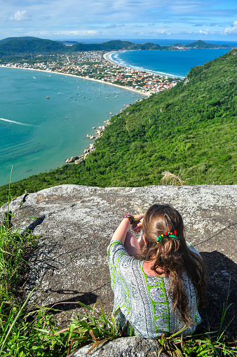 Young woman sitting on a stone looking at Canto Grande and Mariscal Beach from the top of Morro do Macaco Viewpoint. Beautiful sunny day in Bombinhas, Santa Catarina, Brazil