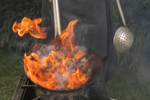 Unrecognizable person flaming meat in a garden using a portable stove and a pan
