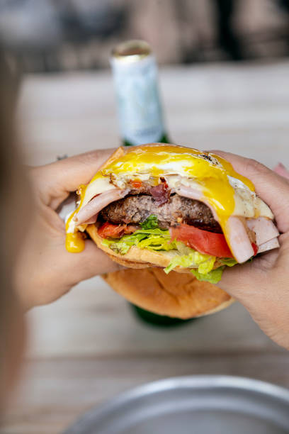 istock Cheeseburger with ham, fried egg, lettuce, tomato and onion, bitten by a person holding it in his hands. 1414147212