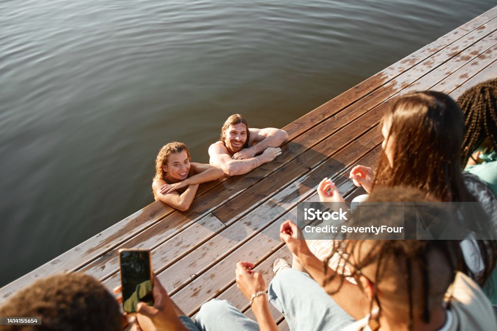Friends Having Fun by Lake High angle view of young couple swimming in lake with group of friends watching from pier and filming video for social media Group Of People Stock Photo