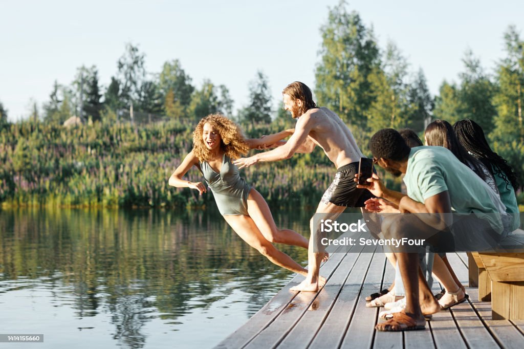 Young People Having Fun at Lake Full length shot of friends having fun at river with playful young man pushing girl in water People Stock Photo