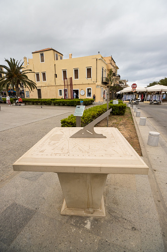 Sundial near The Square of the Unknown Soldier in Rethymnon Town on Crete, Greece, with people and businesses in the background.