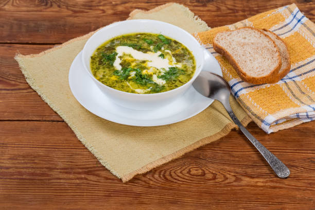 Serving of the sorrel soup, also known as green borscht with chopped hard-boiled egg and sour cream in a white bowl, slices of bread on a napkin on an old rustic table