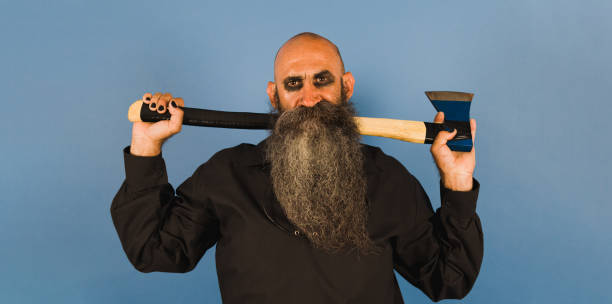istock a long bearded man dressed in balck shirt holding an axe behind his back while looking at camera ahead a blue background 1414182444