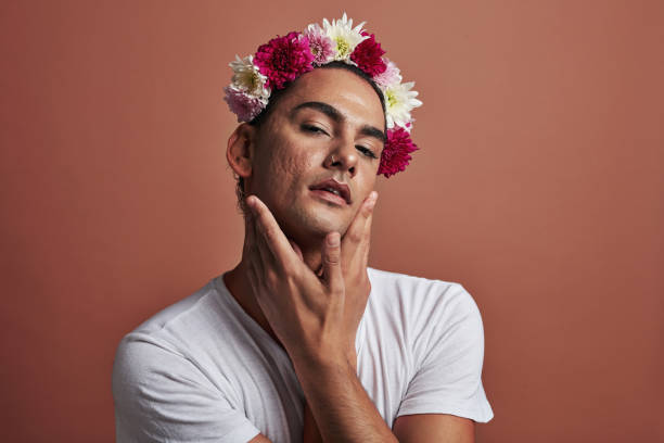 LGBTQ queer, gay or bisexual young man looking confident in a portrait. A beautiful, handsome and attractive gender neutral, androgynous person posing while wearing a floral flower crown An LGBTQ queer, gay or bisexual young man looking confident at the camera portrait. A beautiful, handsome and attractive gender neutral person holding their face with hands and wearing a flower crown gender neutral photos stock pictures, royalty-free photos & images