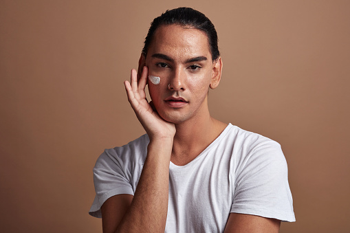 Lgbt male model doing a beauty or skin care routine with anti acne face cream or cosmetic product. Confident gay man moisturizer on cheeks, with brown copyspace background.