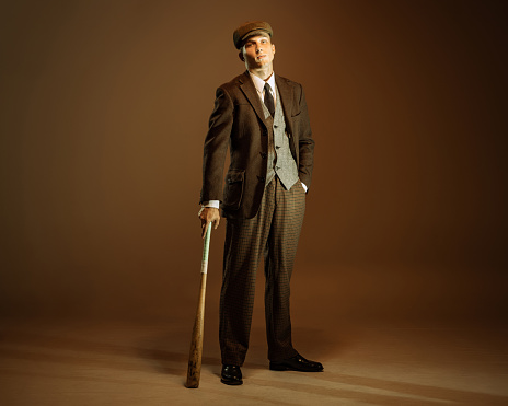 Peaky blinders style. Young man in image of english gangster, businessman wearing suit and cap standing isolated over dark vintage background. Concept of business, personality, emotions, fashion