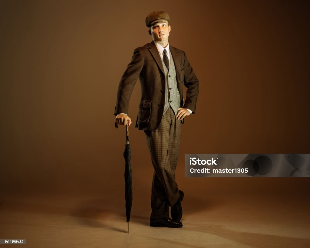Retro style portrait of young man in image of english gangster, businessman wearing suit and cap standing isolated over dark vintage background. Retro style portrait of young man in image of english gangster, businessman wearing suit and cap standing isolated over dark vintage background. Concept of business, personality, emotions, fashion Men Stock Photo