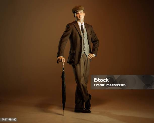 Retro Style Portrait Of Young Man In Image Of English Gangster Businessman Wearing Suit And Cap Standing Isolated Over Dark Vintage Background Stock Photo - Download Image Now