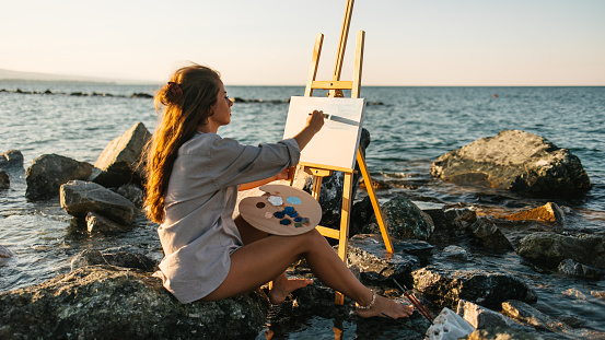 A young artist in beige shirt painting by the sea. Canvas on an easel. Easel placed on the rocks. Legs in the water. Waves hitting the rocks