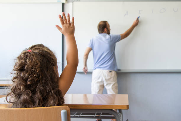 student raising her hand in a class lesson while the teacher explains at school stock photo
