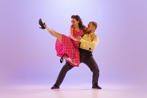 Lindy Hop. Active and emotional couple in colorful retro style costumes dancing incendiary dances isolated on purple background in neon. Actors in motion and action. Concept of art, 60s, 70s culture