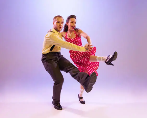 Photo of Emotional bright couple of dancers in colorful retro style attires dancing incendiary dances isolated on purple background in neon light.