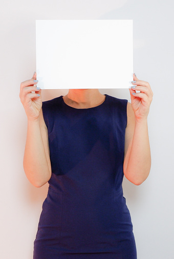 An unrecognizable attractive woman in a blue dress holds a white sheet of paper. Place for your text. Light background.