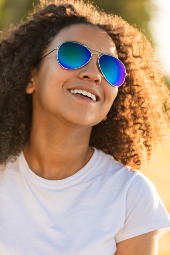 A beautiful and happy mixed race African American female girl child teenager young woman in sunshine wearing blue reflective aviator sunglasses and smiling with perfect teeth