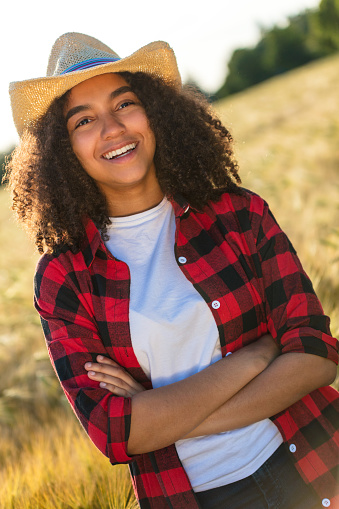Portrait of beautiful and happy mixed race African American female girl child wearing straw cowboy hat and plaid shirt and white t-shirt, smiling with perfect teeth in a wheat or barley field at sunset