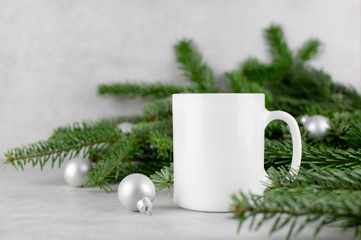Blank white mug mockup with christmas tree branches and silver balls on light concrete stone background. Holiday composition. Side view. Copy space.