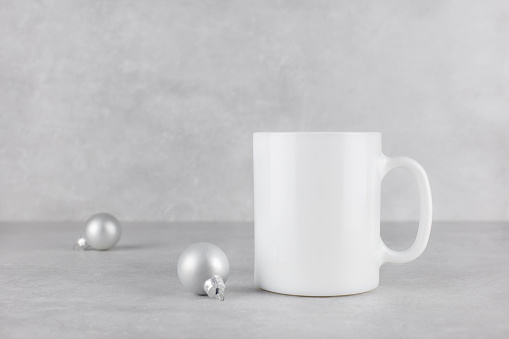 Blank white mug mockup with christmas silver balls on light concrete stone background. Holiday composition. Side view. Copy space.