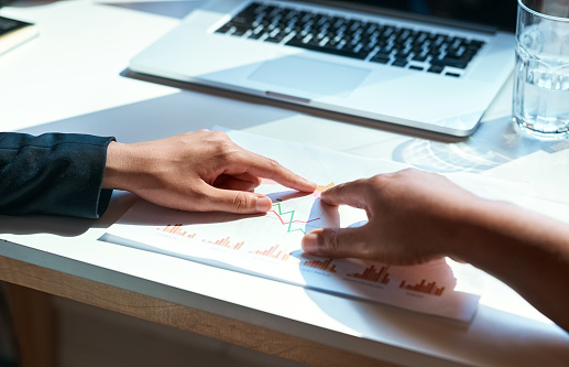 Hands pointing to the market chart on a paper with their fingers together in a team business meeting. Cropped close up of diverse corporate businesspeople working on a sales strategy with a laptop