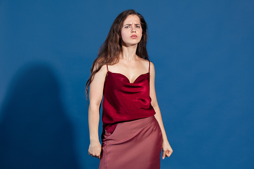 Portrait of young emotive woman posing with serious look isolated over blue studio background. Stylish deep red dress. Concept of beauty, lifestyle, youth, emotions, facial expression, ad