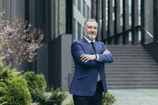 Portrait of successful director and business owner, man with crossed arms smiling outside office building, businessman investor in business suit confident