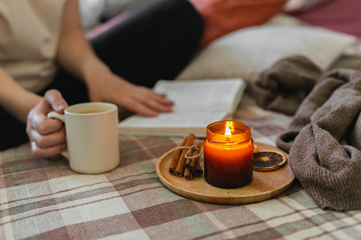 Young woman reading book while sitting in lotus pose on bed in cozy bedroom, spending leisure time at home. Burning candle. Cozy lifestyle, hygge concept