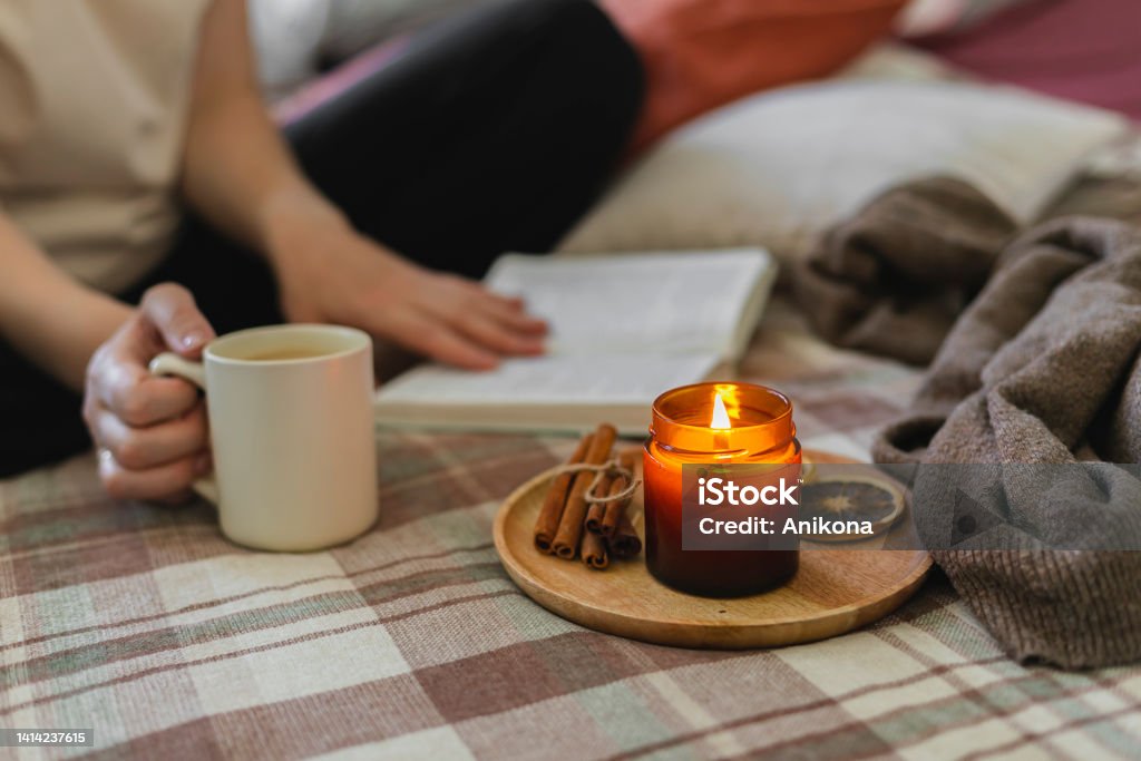Young woman reading book while sitting in lotus pose on bed in cozy bedroom, spending leisure time at home. Young woman reading book while sitting in lotus pose on bed in cozy bedroom, spending leisure time at home. Burning candle. Cozy lifestyle, hygge concept Hygge Stock Photo