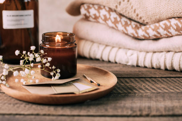 vela encendida en un pequeño frasco de vidrio ámbar, flores de gypsophila y suéteres de costura de punto apilados. estilo de vida acogedor, concepto hygge - acogedor fotografías e imágenes de stock