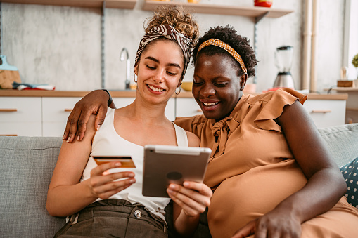 Young beautiful black pregnant woman and her partner sitting on the sofa in the living room and shopping online using tablet.