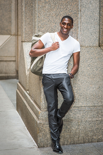 Street Fashion. Young African American man wearing white V neck T shirt, black pants, leather shoes, carrying shoulder bag, standing against column outside in New York City, smiling, looking at you.