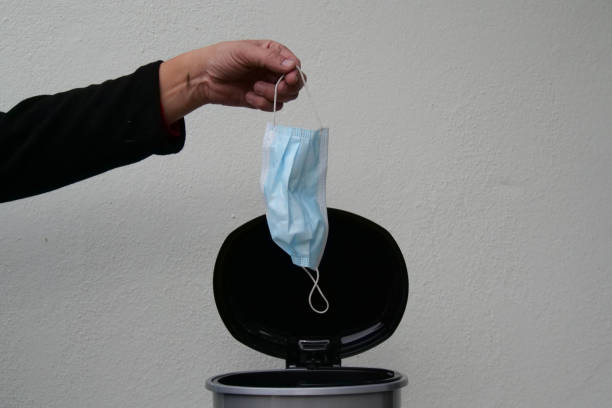 istock Hand holding light blue disposable face mask before throwing it away in a black bin signifying the end of the corona virus pandemic. 1414249804