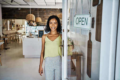 Entrepreneur, startup and business owner welcoming people to the opening of her new cafe and shop. Friendly and smiling woman standing and waiting by the doorway and entrance of her coffee shop