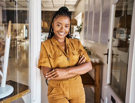 Startup and small business entrepreneur welcoming you to the opening of her cafe or coffee shop. Portrait of cheerful and smiling owner standing in the entrance or doorway with her arms crossed