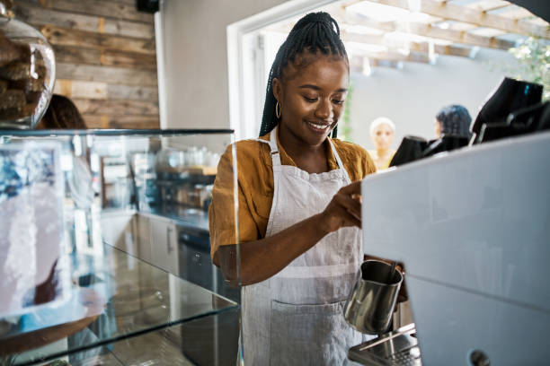 istock Barista making cappuccino at a coffee shop or bar using a machine on a morning during the weekend. African American female waitress wearing an apron preparing a hot beverage at a cafe 1414263295