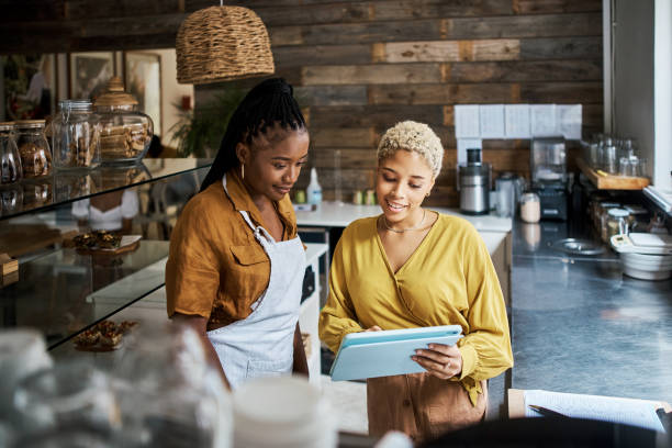 Coffee shop manager, owner and supervisor using a tablet to train a waitress in her shop. Workers talking while checking online orders and using a mobile app for social media ads in a cafe stock photo