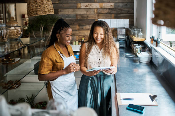 Two restaurant workers planning on a digital tablet in a small coffee shop. Happy female colleagues reviewing plans and reports in a cafe and doing digital online stocktake and orders