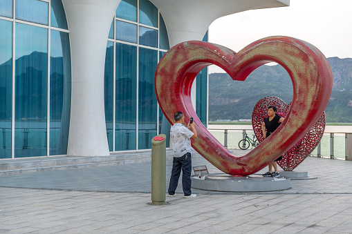 Wenzhou, China - July 16, 2022: A man using a mobile phone to snap the picture of a woman posing in front of a red heart-shaped artwork. There is a river and mountain in the background.