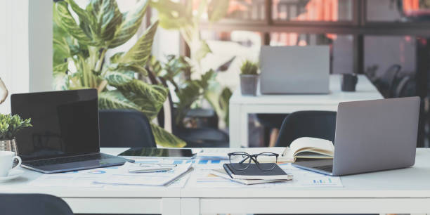 istock Laptop Computer, notebook, and eyeglasses sitting on a desk in a large open plan office space after working hours 1414266137
