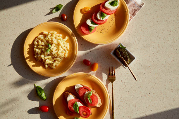 istock Mediterranean pasta fusilli, caprese salad - tomatoes, mozzarella, basil and pesto. Bread appetizer with caprese salad on orange plates under hard shadows. Top view food flat lay. Copy space. 1414266514