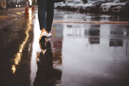 Unidentified woman walking a long the street while raining, wet concrete road from rain storm. Woman stepping on the downtown street at night, close up on foots.