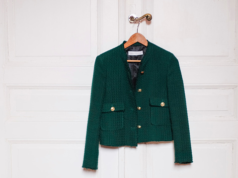 Stylish women's trendy green jacket with gold large buttons hangs on a wooden coat hanger over the doorknob of an antique white door with little copy space, close-up side view. The concept of fashion accessories.