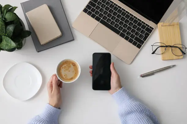 Photo of Top view of woman using smartphone and cup with coffee at white office table