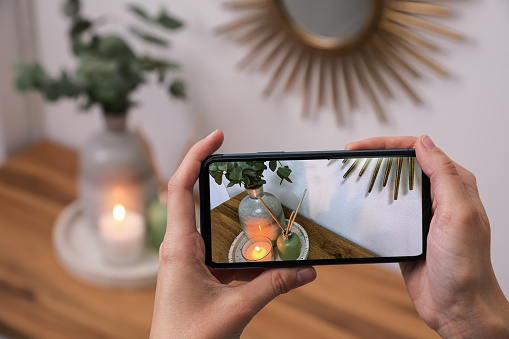 Woman taking photo of stylish interior accessories on wooden table with smartphone, closeup