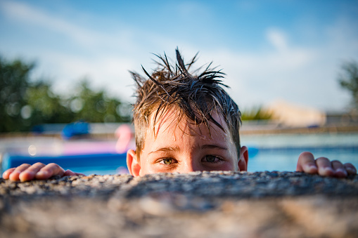 A young boy peaks over the edge of a swimming pool. Just half of his face is visible. His fingers are just on the edge. He is has playful look in his eyes.