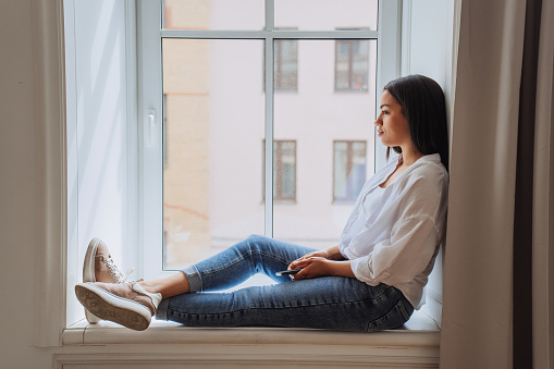 Sad Brazilian young woman sitting on windowsill in white shirt and blue jeans frustrated after divorce, thinking about future holding phone at home, sunny day. Pretty African American girl in troubles