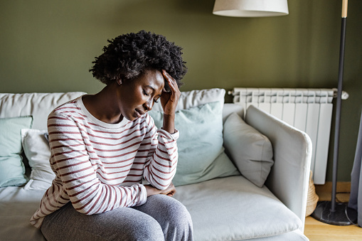 Young African American woman sitting on the couch at home. She is holding her head like she is having pains.