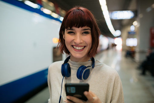 Portrait of a happy young adult walking through the subway station , using a smart phone and searching for her train while looking at the camera