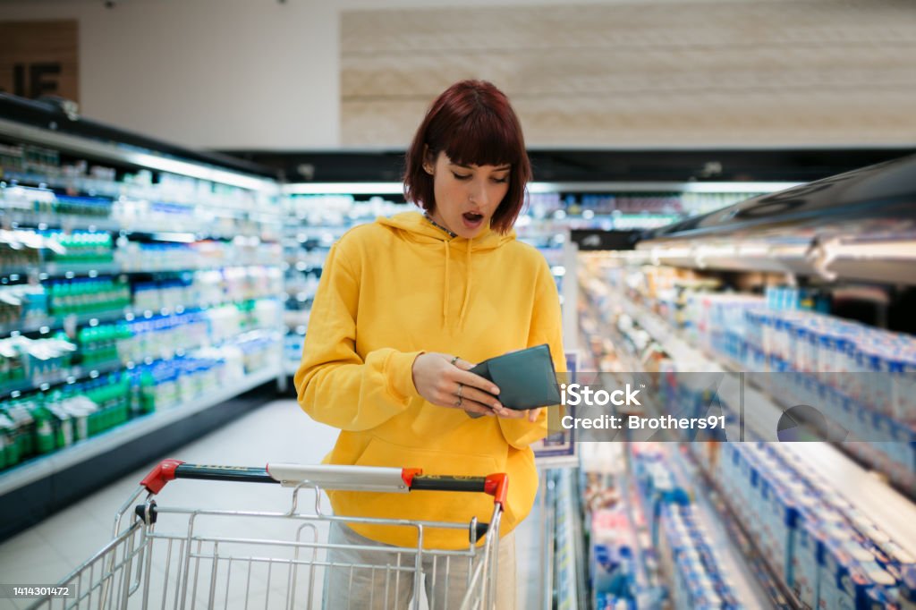 Shocked by the prices Young adult woman with red hair buying groceries in a local supermarket, expressing a state of shock on her face after seeing the prices of food on the shelfs, looking at her wallet Empty Wallet Stock Photo